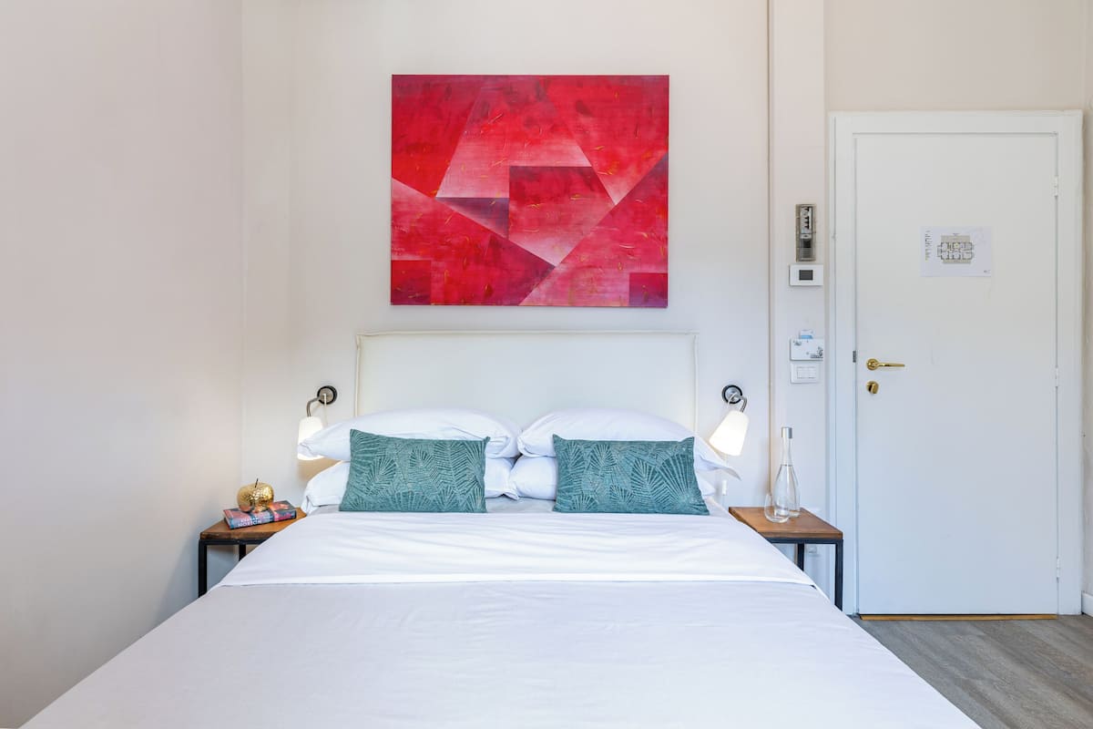 A neatly made bed with white linens and two decorative pillows, a wall-mounted abstract painting, a bedside table with a lamp, and a door with a lock.