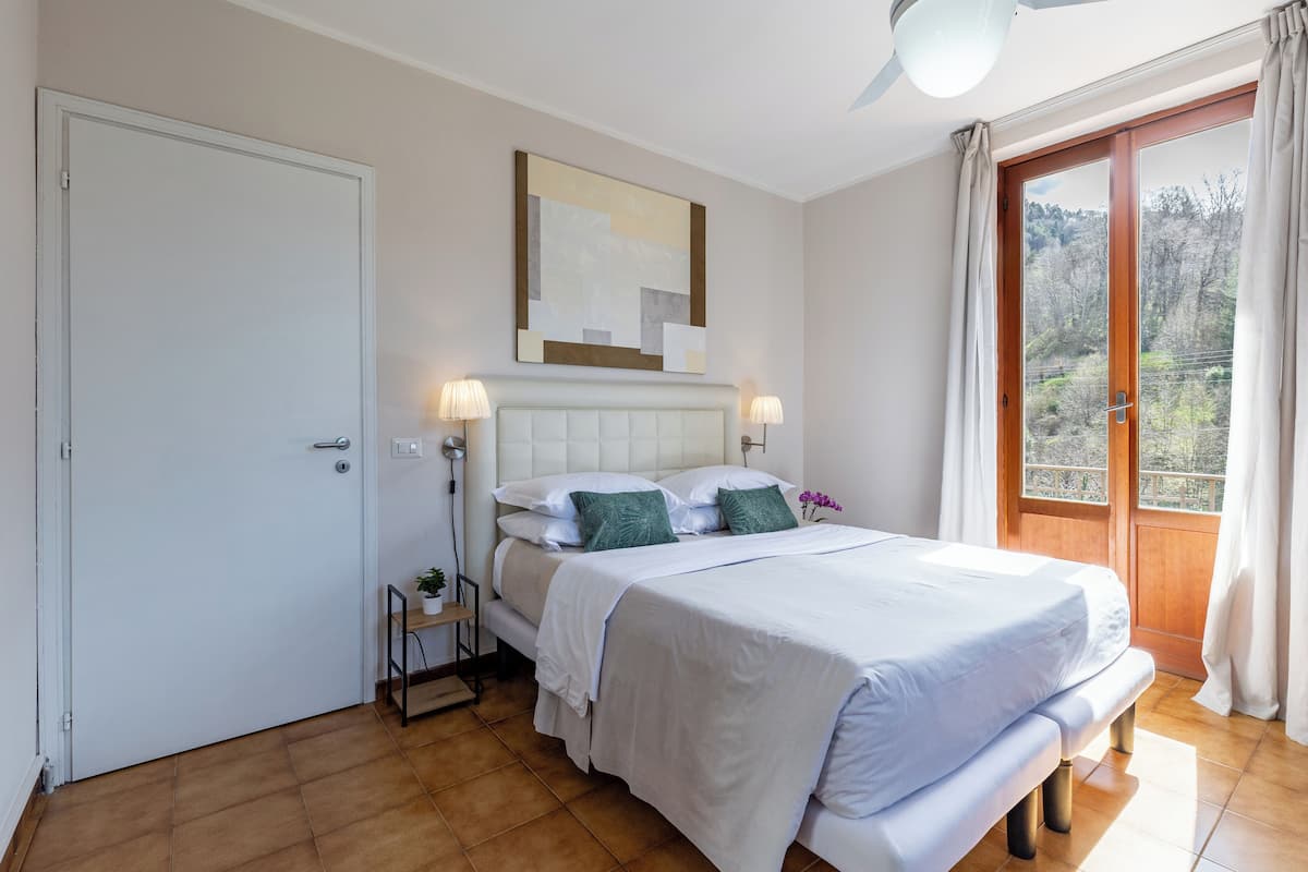 A bedroom with a bed, bedside lamps, a nightstand, a window with curtains, and a view of trees.