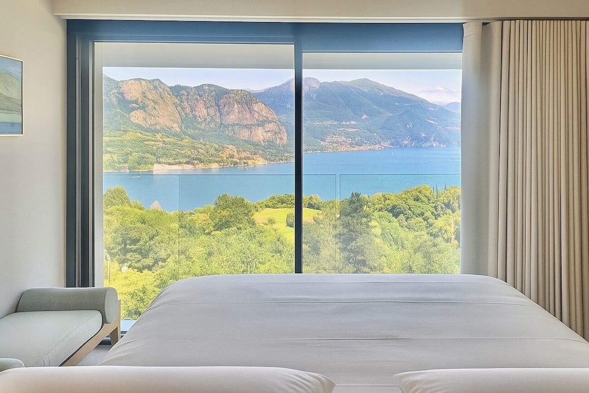 A bedroom with a large bed, a chair, and a view of mountains and a lake.