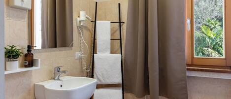 A bathroom with a white sink, a towel rack, a shower, and a window with a view of greenery.