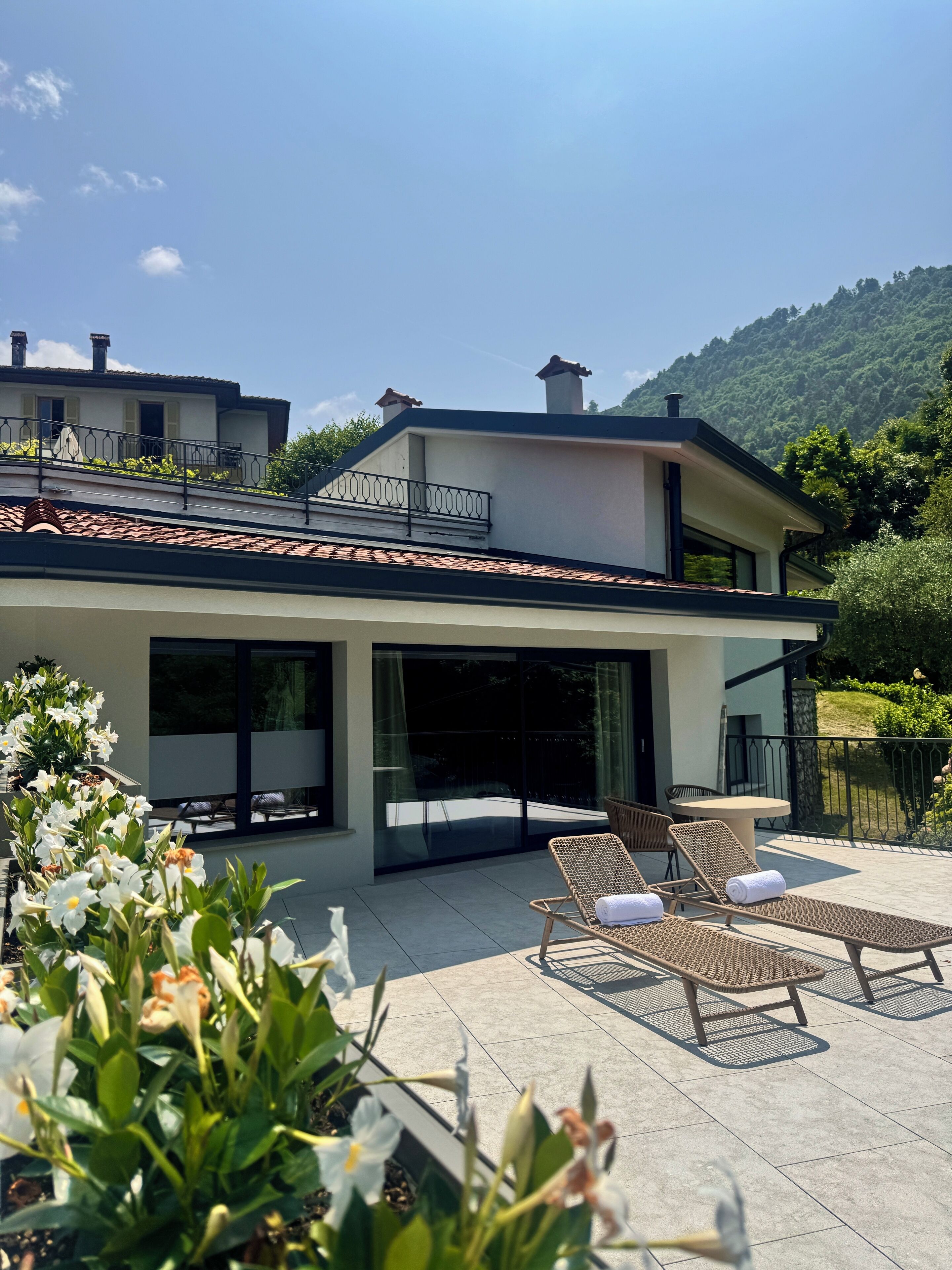 A modern house with a large glass door, outdoor seating, and a garden with flowers.