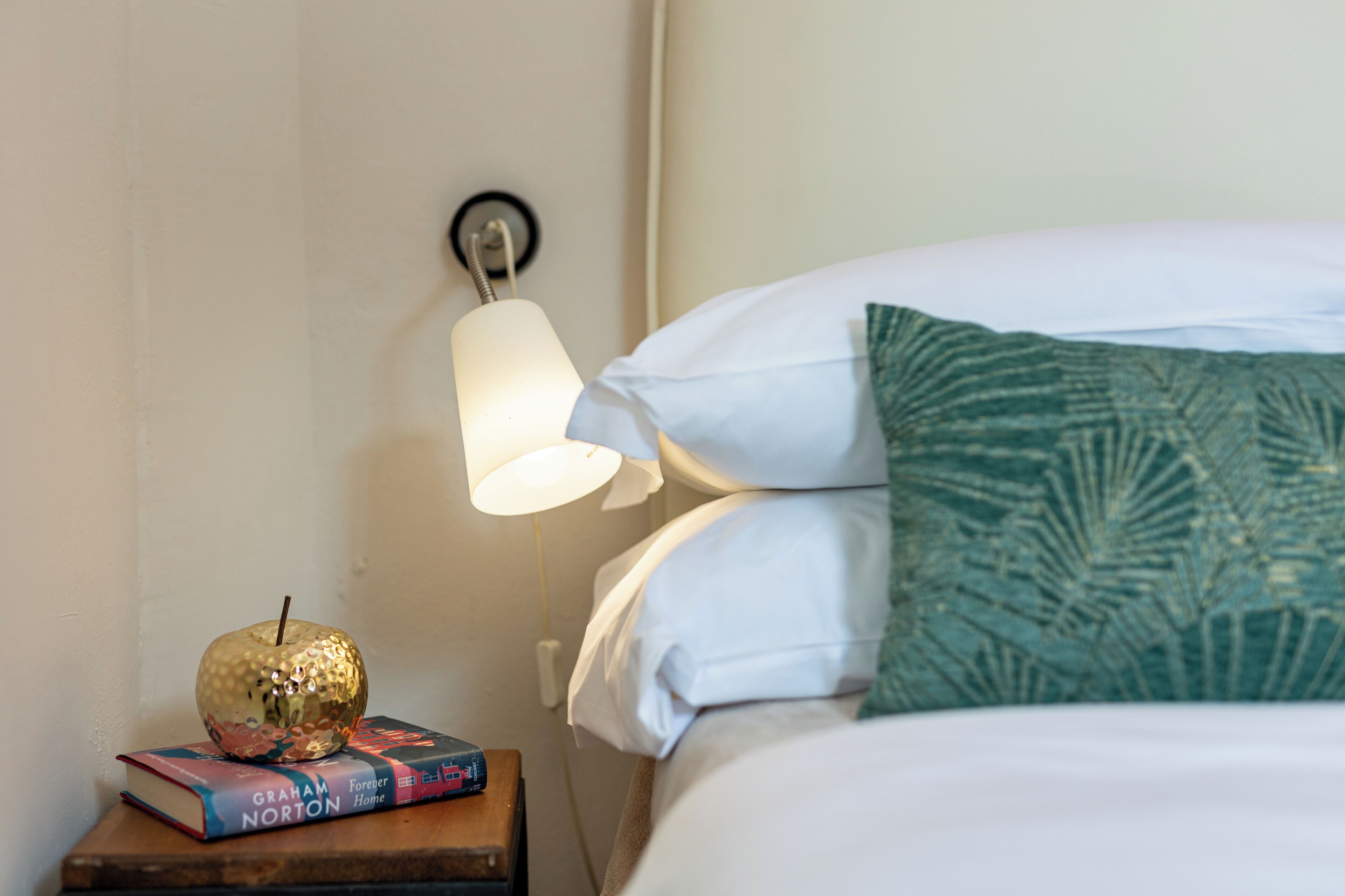 Una cama bien hecha con almohadas blancas y verdes, una mesita de noche con libros y una manzana decorativa, y una lámpara de pared.