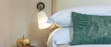 Una cama bien hecha con almohadas blancas y verdes, una mesita de noche con libros y una manzana decorativa, y una lámpara de pared.