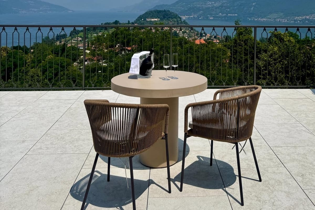 A terrace with a round table and two wicker chairs, overlooking a mountainous landscape with a view of the sea.