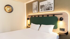 Una cama bien hecha con ropa blanca, un cabecero tapizado en verde y dos lámparas de lectura fijadas a la pared.