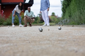 A close-up of a billiards table with balls in motion, a person in the background, and a red building with greenery around. - Campanile NATURE - Orange (Orange)