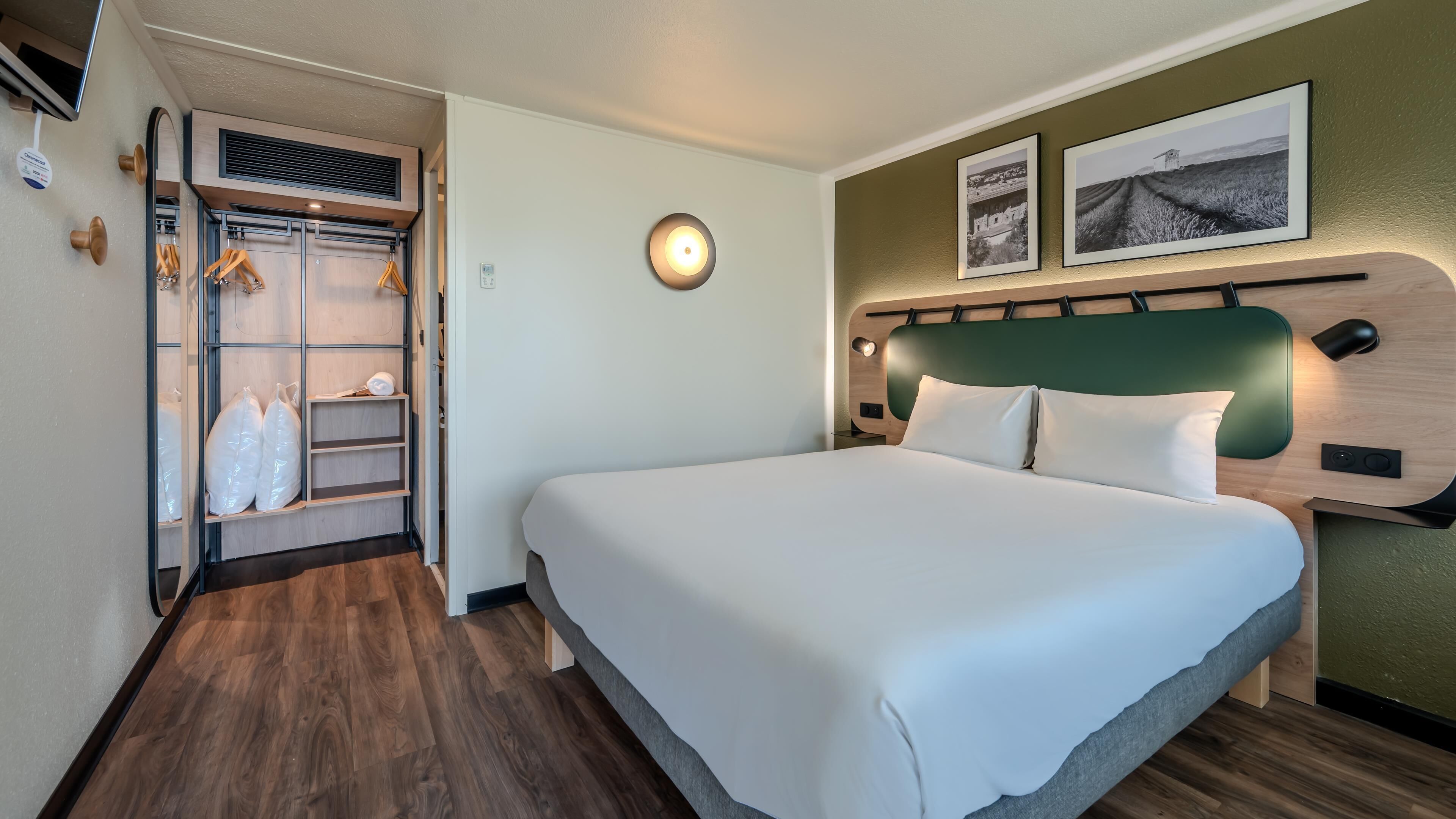 A hotel room with a bed, a closet with hanging clothes, a round wall light, and two framed pictures on the wall.