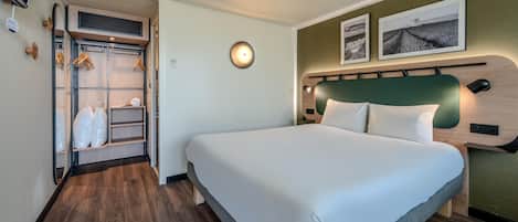Quarto de hotel com cama, armário com roupas penduradas, luminária redonda na parede e duas imagens emolduradas.