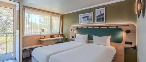 A hotel room with two beds, a desk, a chair, a window with curtains, and two framed pictures on the wall.