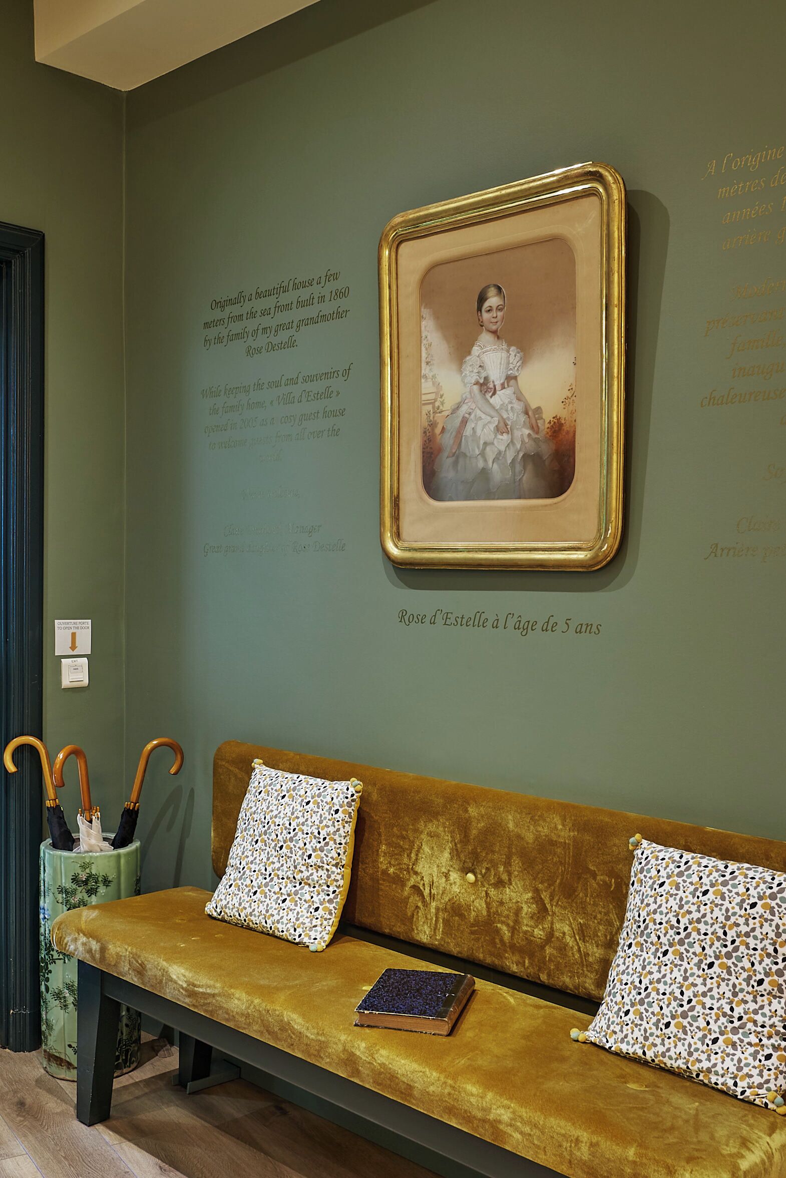 A room with a green wall, a framed portrait, a bench with patterned cushions, and a small table with a book.