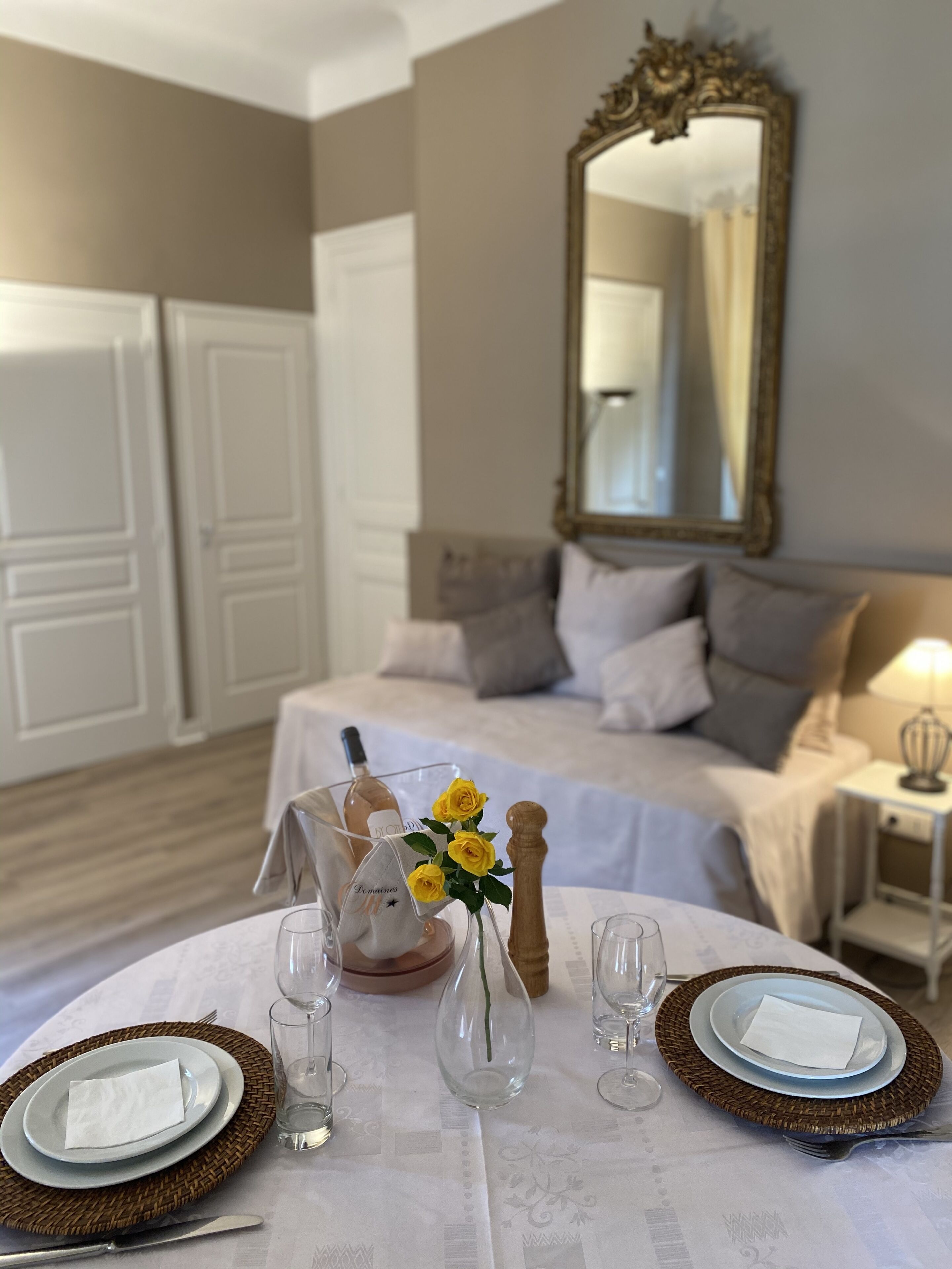 Una mesa de comedor bien puesta con platos, cubiertos y un florero con flores amarillas. En el fondo, una cama con almohadas y una mesita de noche con una lámpara.