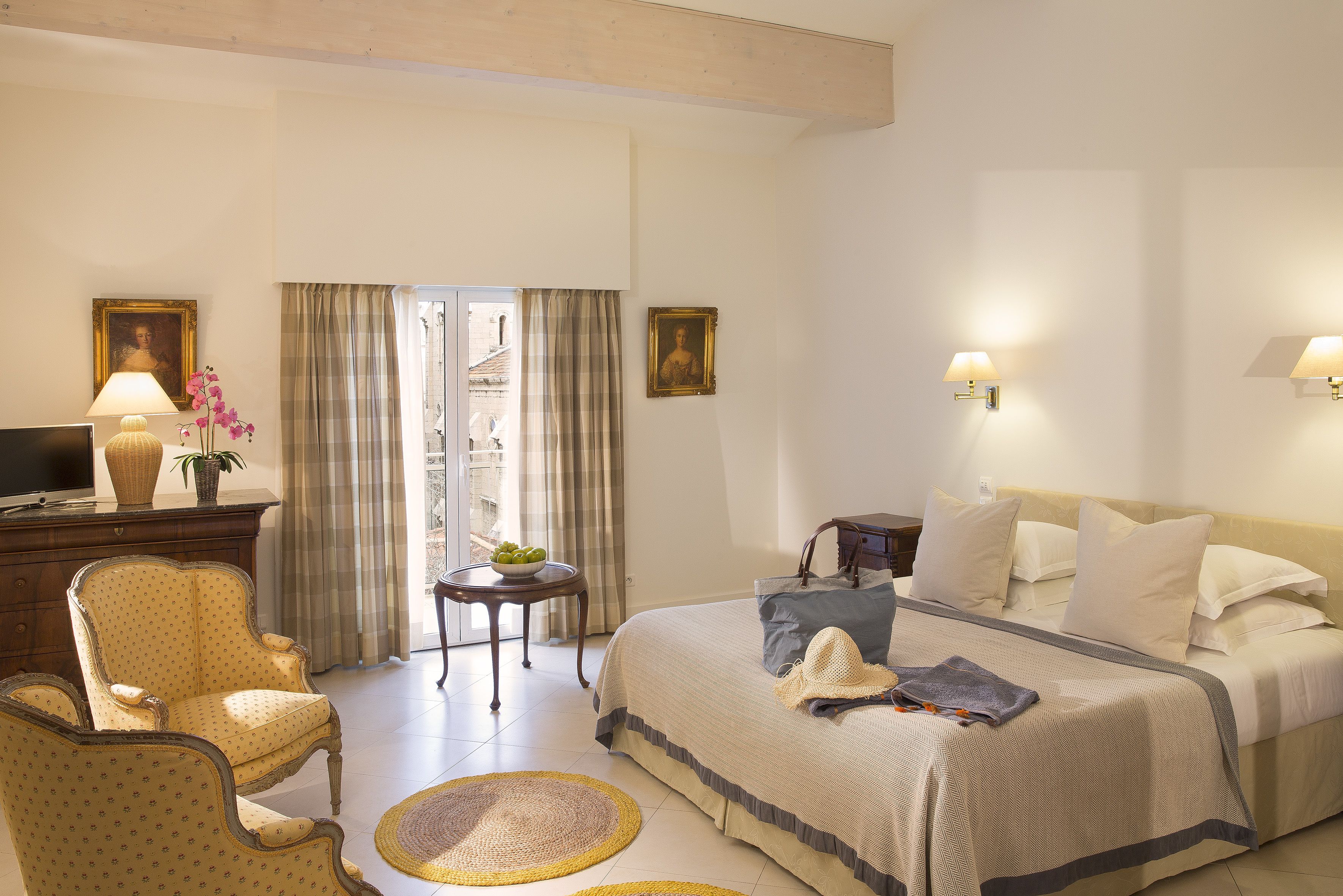 A hotel room with a bed, a chair, a desk with a lamp, a small table with a plant, and a window with curtains.