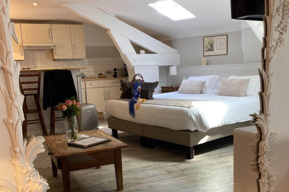 A modern, well-lit room with a bed, a small table with a vase, and a kitchenette in the background.