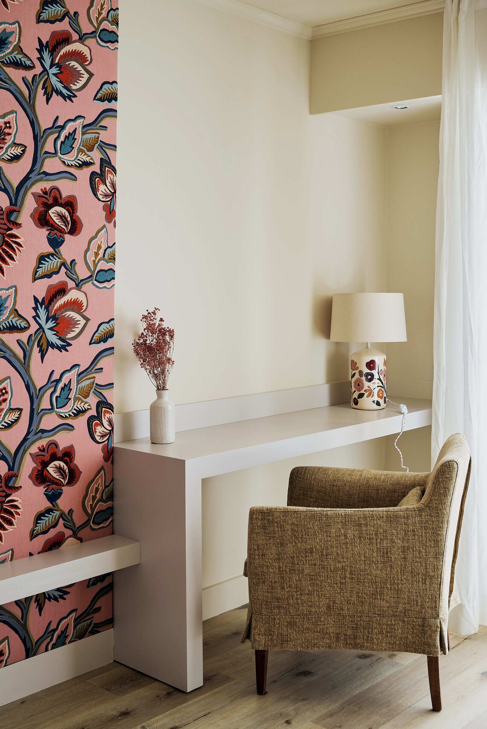 A room with a white desk, a chair, a lamp, and a floral-patterned wall.