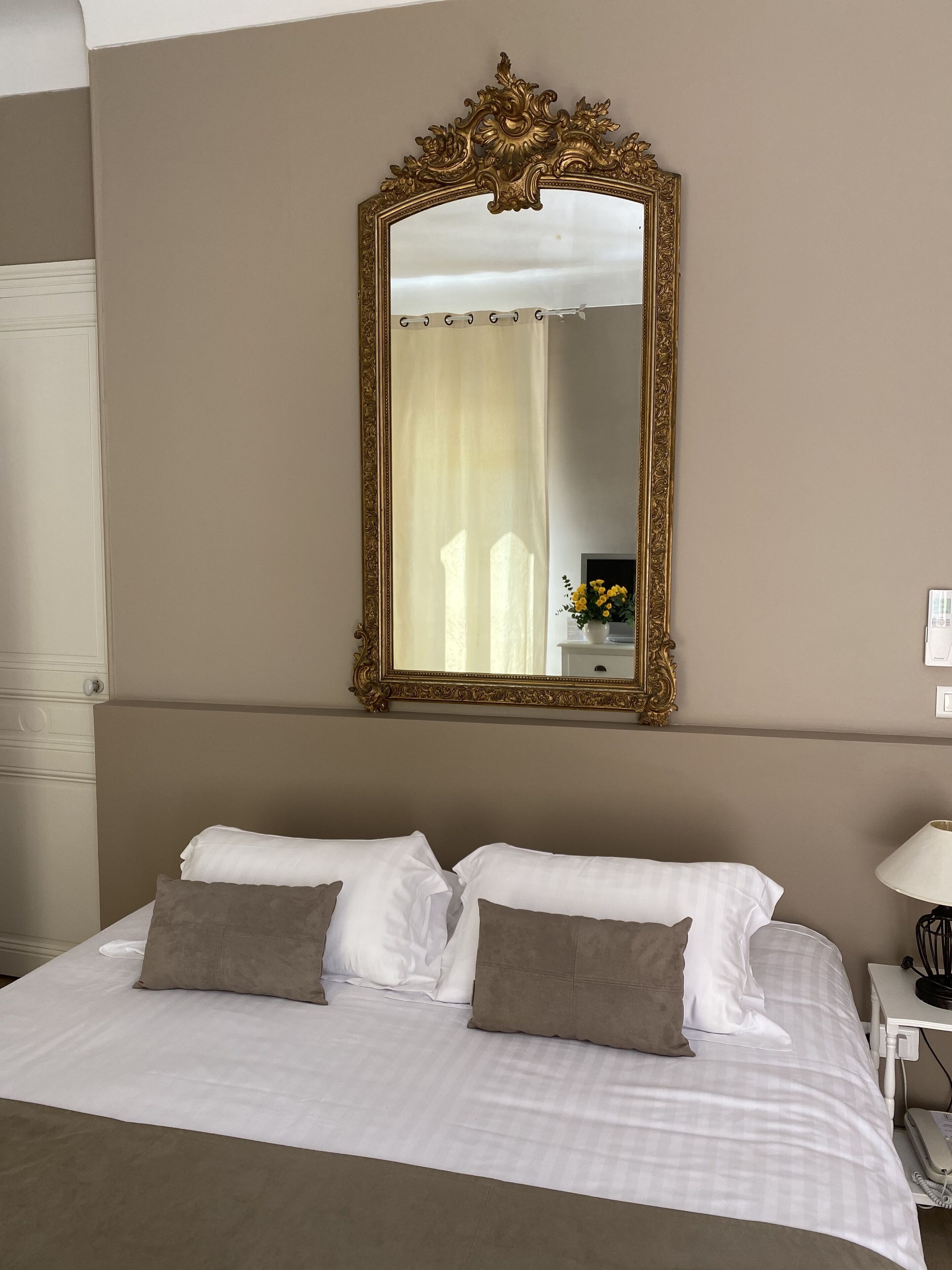 A hotel room with a large ornate mirror, a bed with white and grey bedding, a bedside table with a lamp, and a white wardrobe.