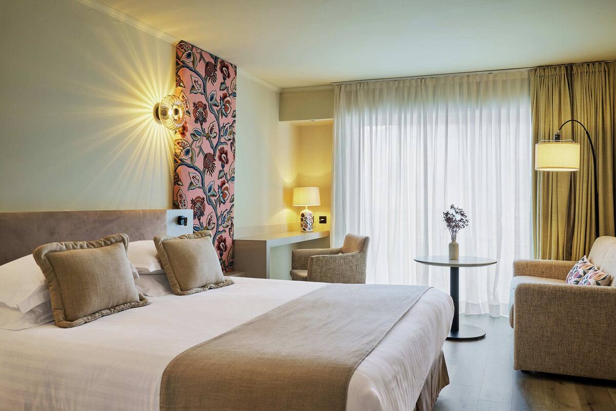 A hotel room with a large bed, two armchairs, a small table with a vase, and a decorative wall panel.