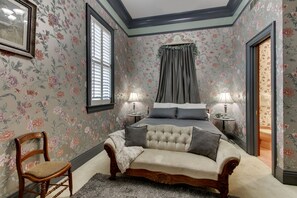 A bedroom with floral wallpaper, a bed with grey bedding, two bedside tables with lamps, and a wooden chair.