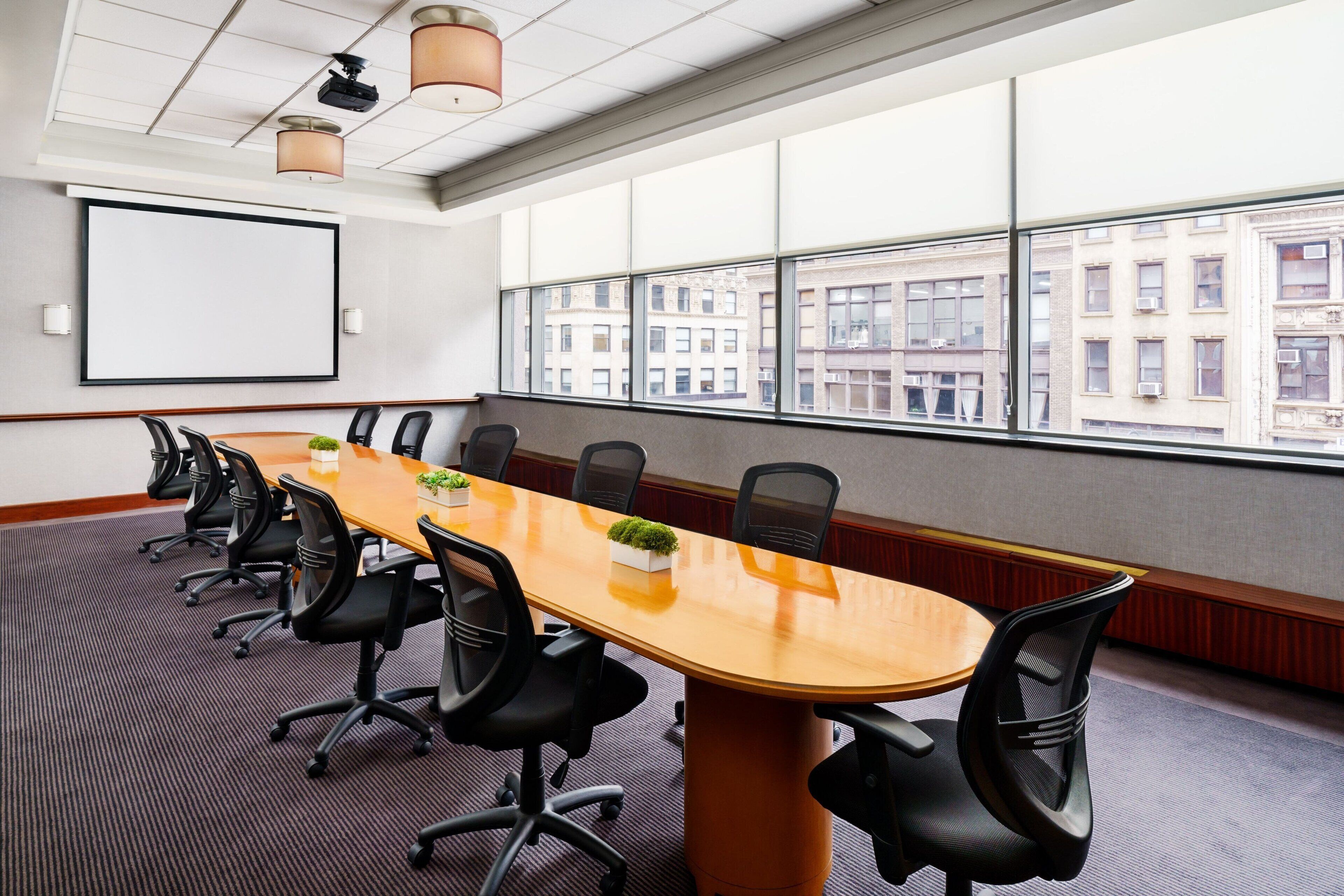 A conference room with a long table, black chairs, a projector screen, and large windows with a city view.