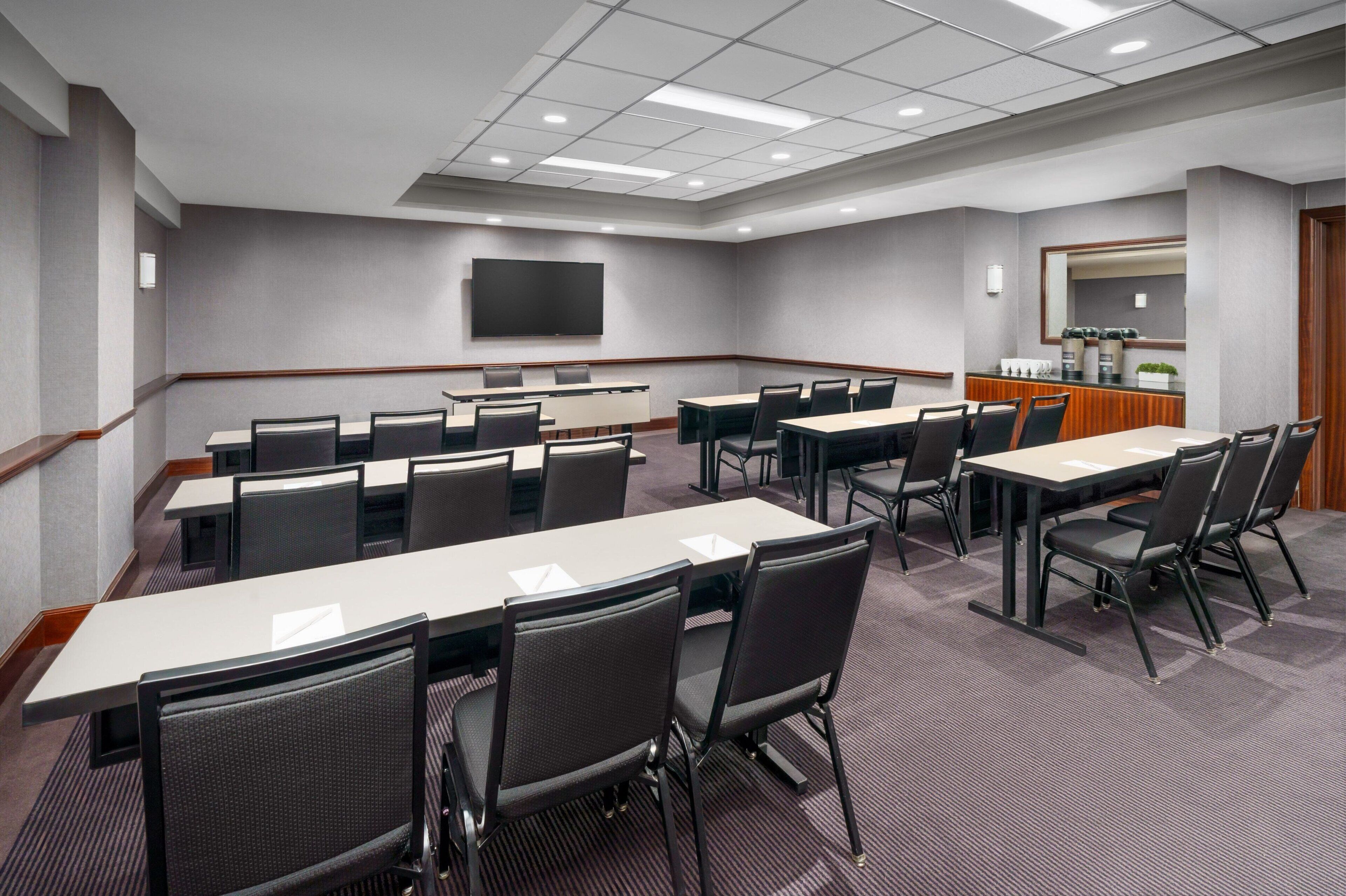 Une salle de conférence équipée de plusieurs tables et chaises, d’un téléviseur à écran plat fixé au mur et d’un petit coin cuisine.