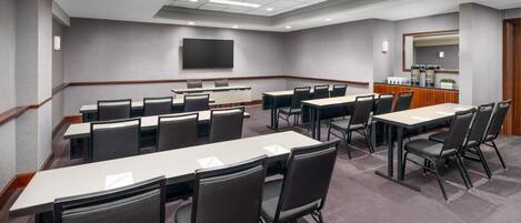 Une salle de conférence équipée de plusieurs tables et chaises, d’un téléviseur à écran plat fixé au mur et d’un petit coin cuisine.