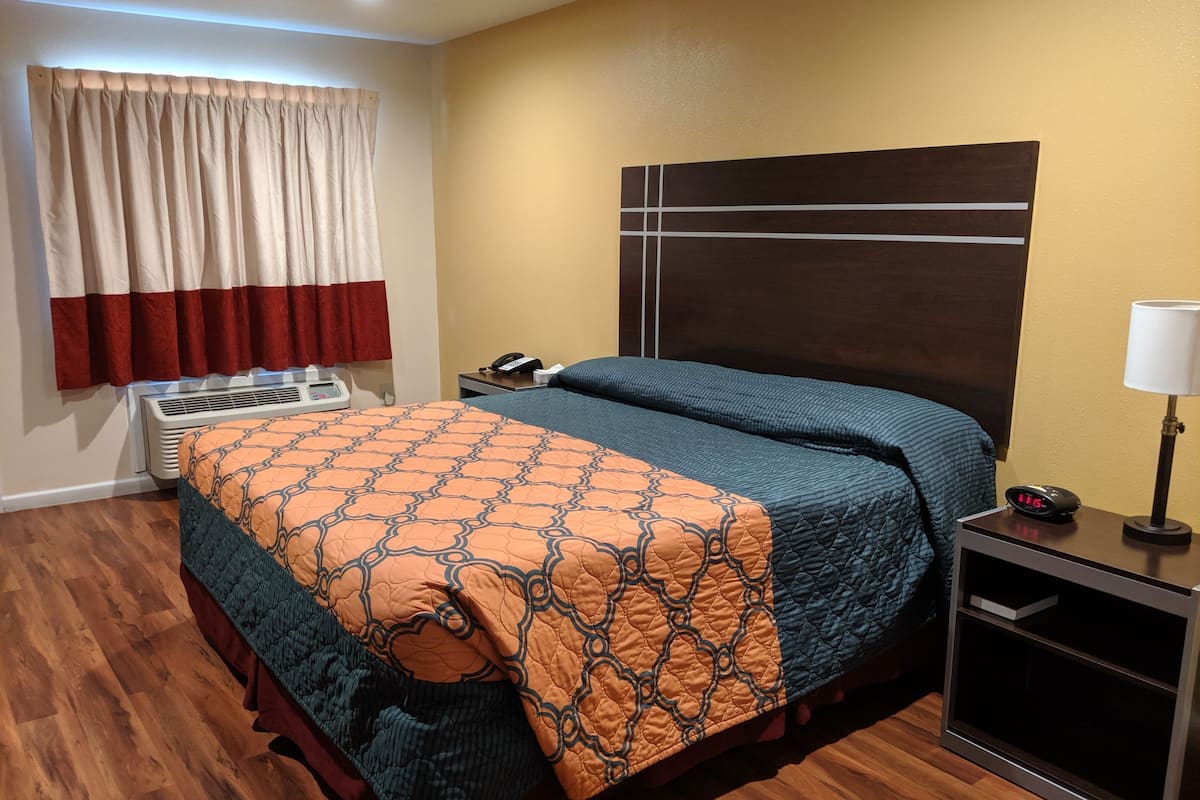 A hotel room with a bed, a nightstand, a lamp, a clock, and a wall-mounted air conditioning unit.