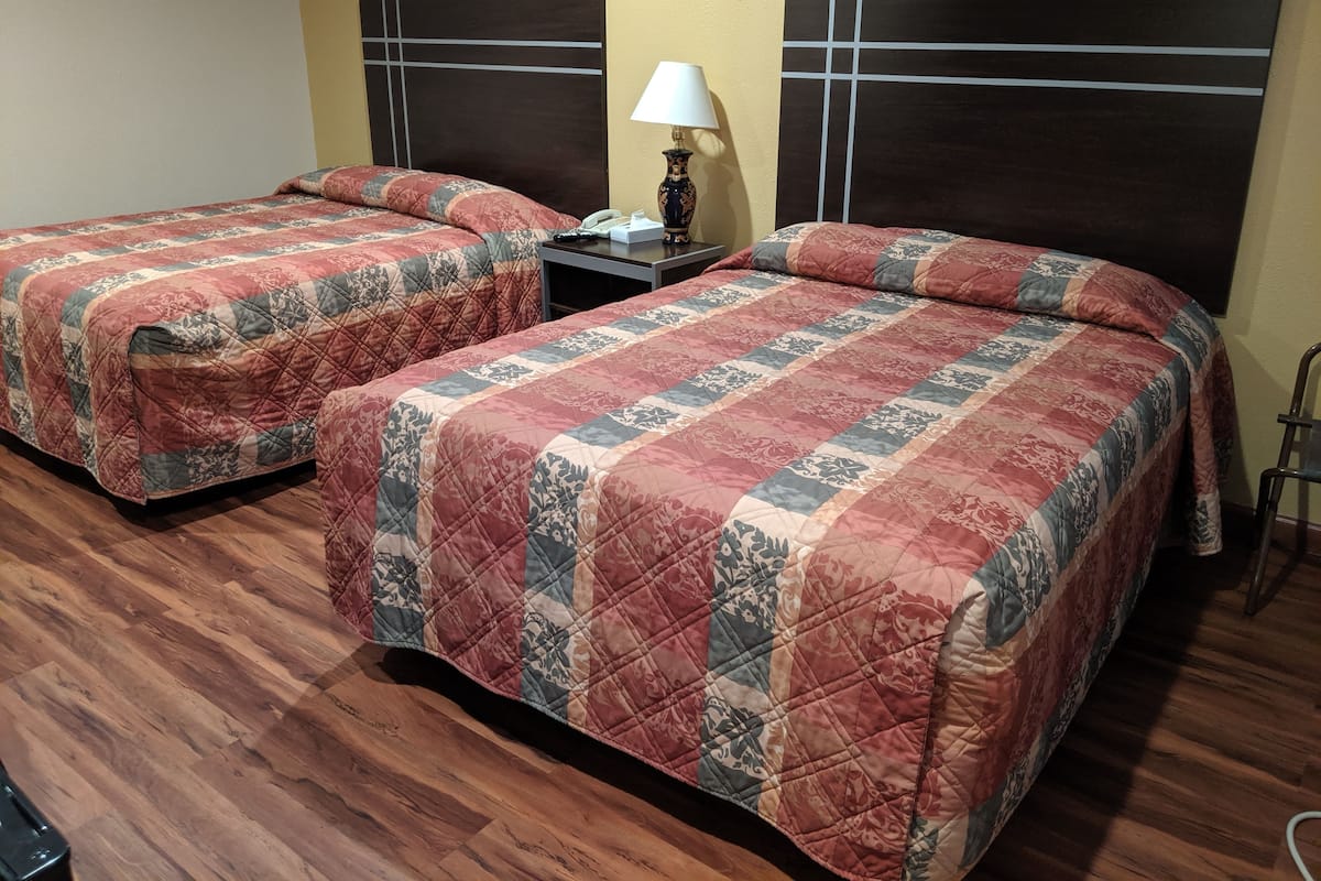 Two beds with matching quilts, a nightstand with a lamp, and a wooden floor.