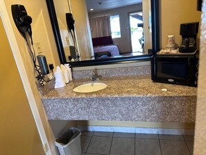 A hotel bathroom with a granite countertop, a sink, a mirror, a hairdryer, and a coffee maker.