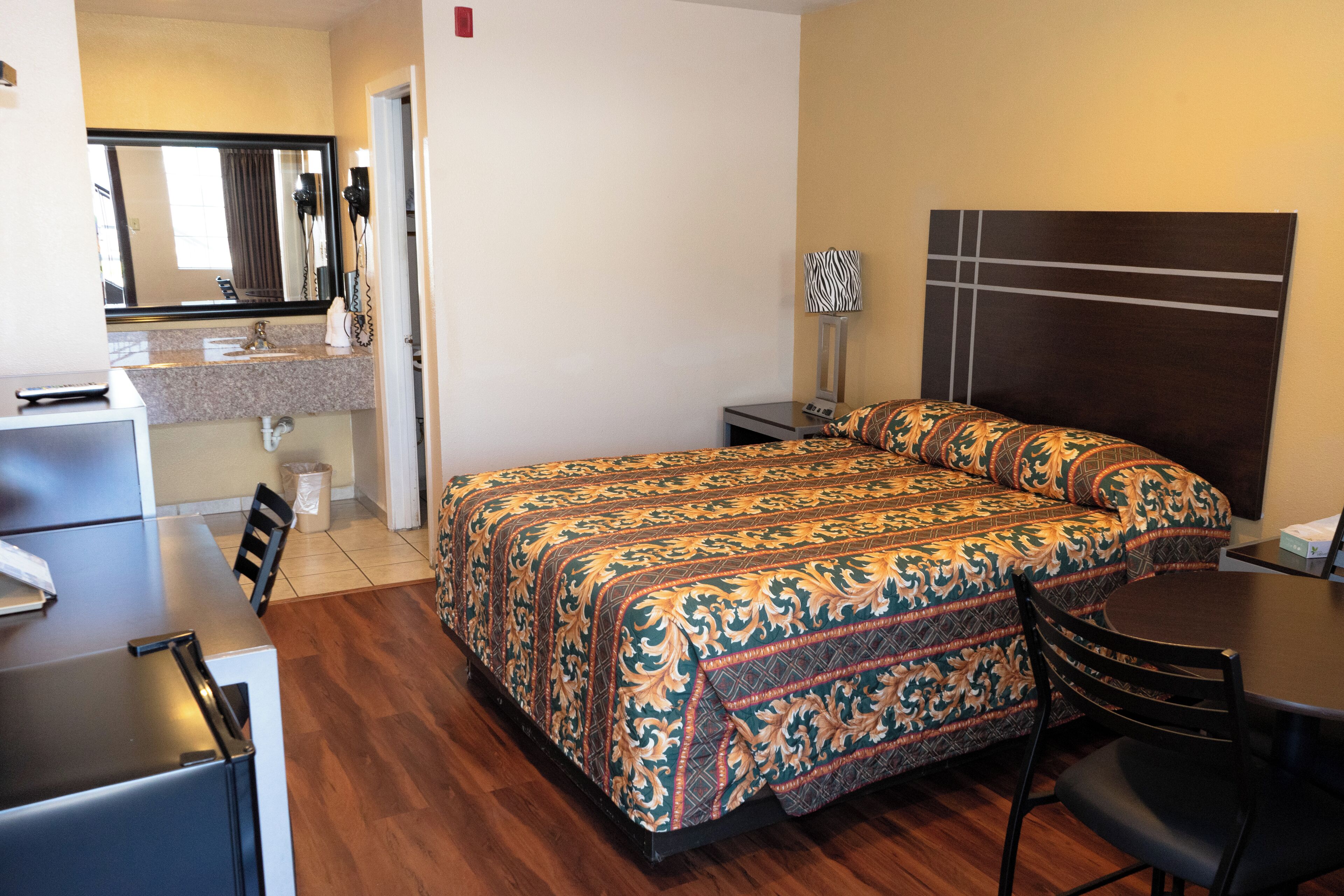 A hotel room with a bed, a desk, a chair, a mirror, and a nightstand with a lamp.