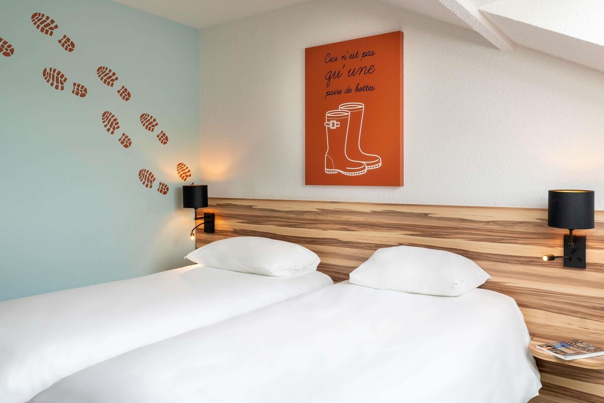 Habitación de hotel con una cama, dos lámparas fijadas a la pared, una pared con huellas y un cartel con botas y texto en francés.