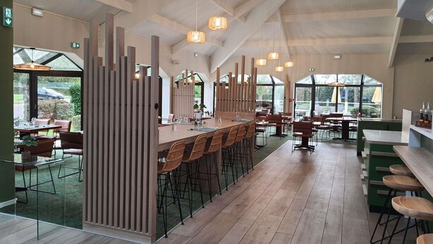 Een ruime eetzaal met hoge plafonds, houten balken en grote ramen met uitzicht op de omgeving.