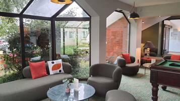 A modern living room with a sofa, armchairs, a round coffee table, and a pool table. Large windows offer views of greenery and a brick wall.