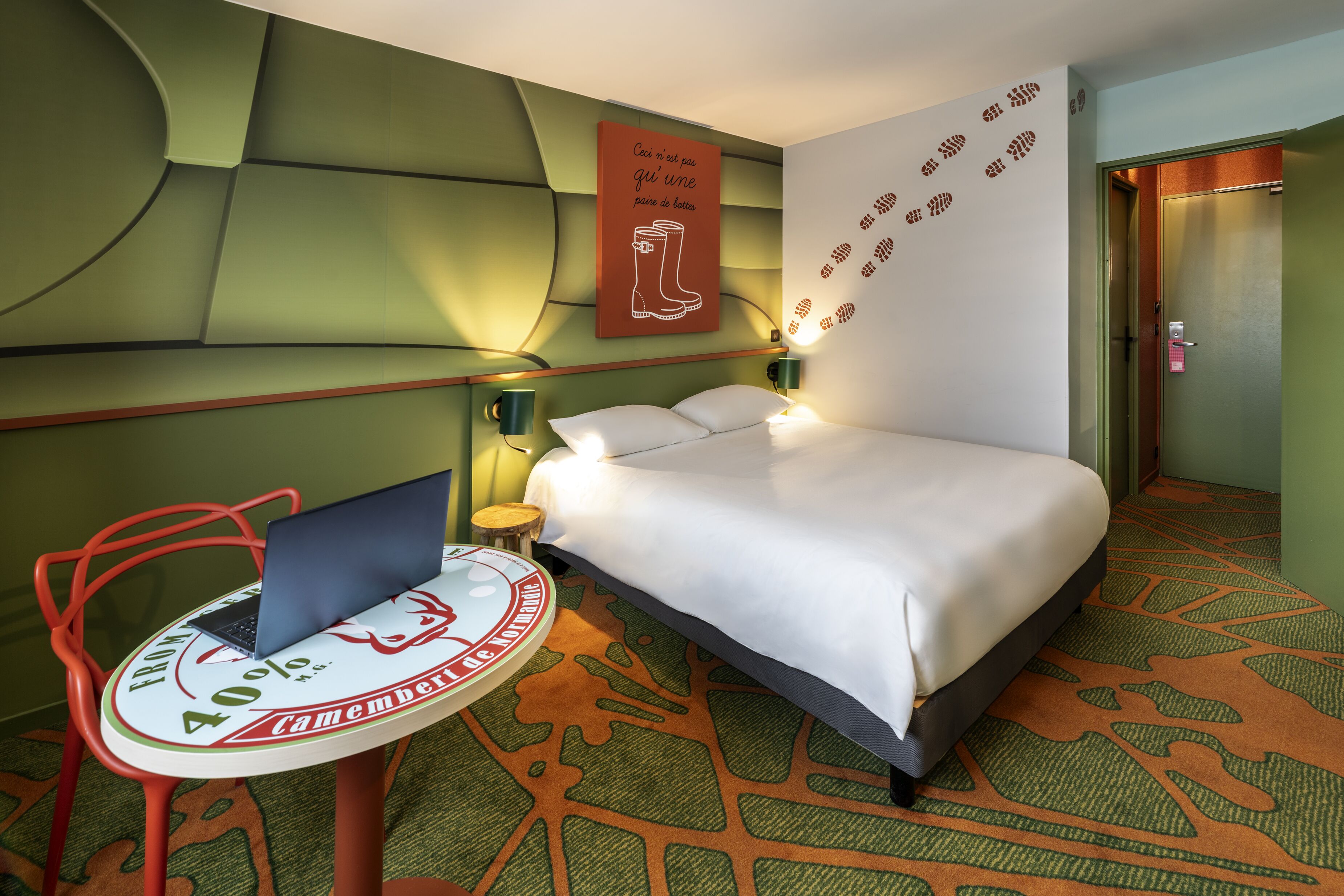 Una habitación de hotel con cama, escritorio con computadora portátil, pared con un cartel y una puerta.