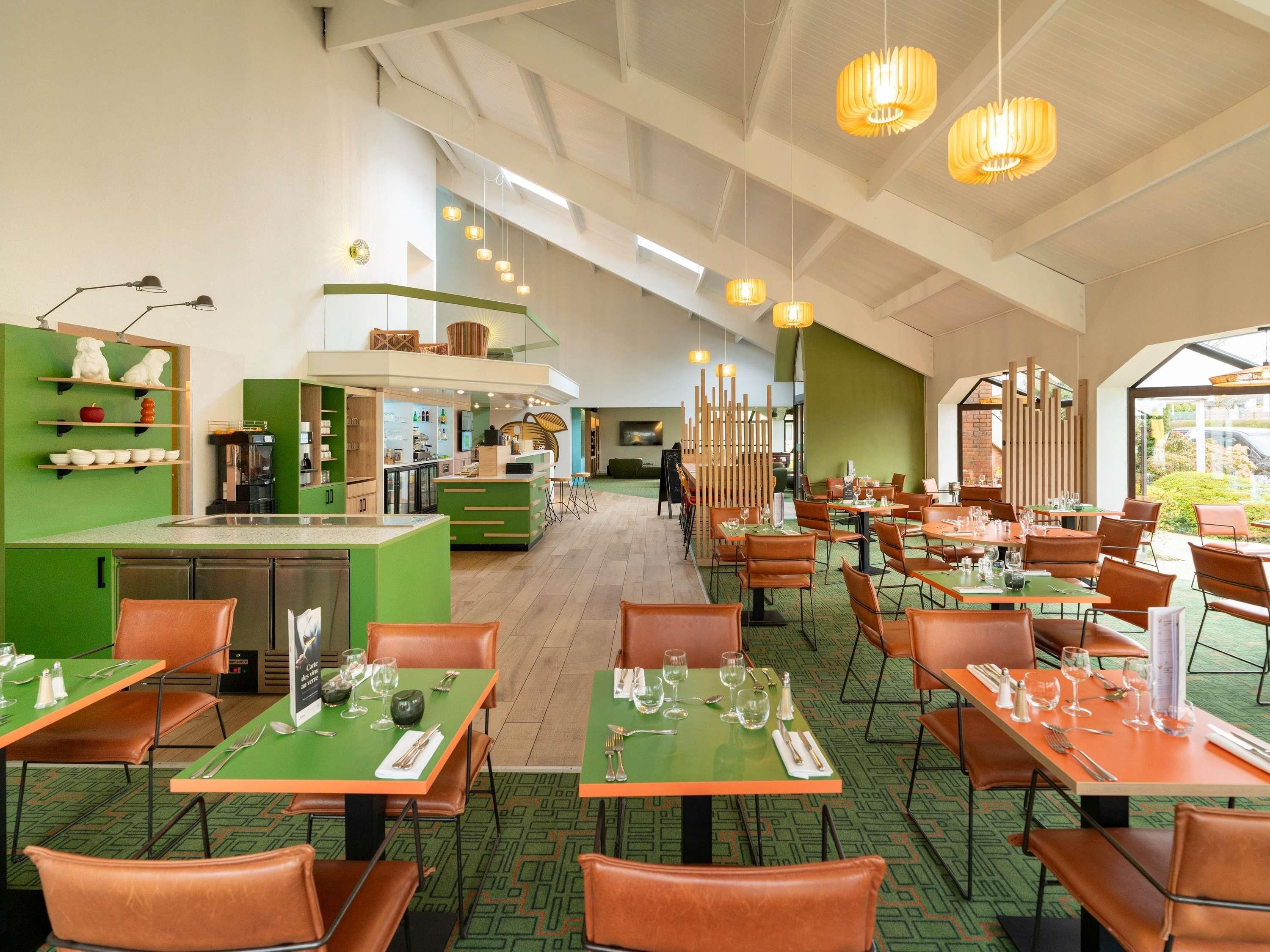 Un restaurante con mesas y sillas verdes, un área de cocina y lámparas colgantes.