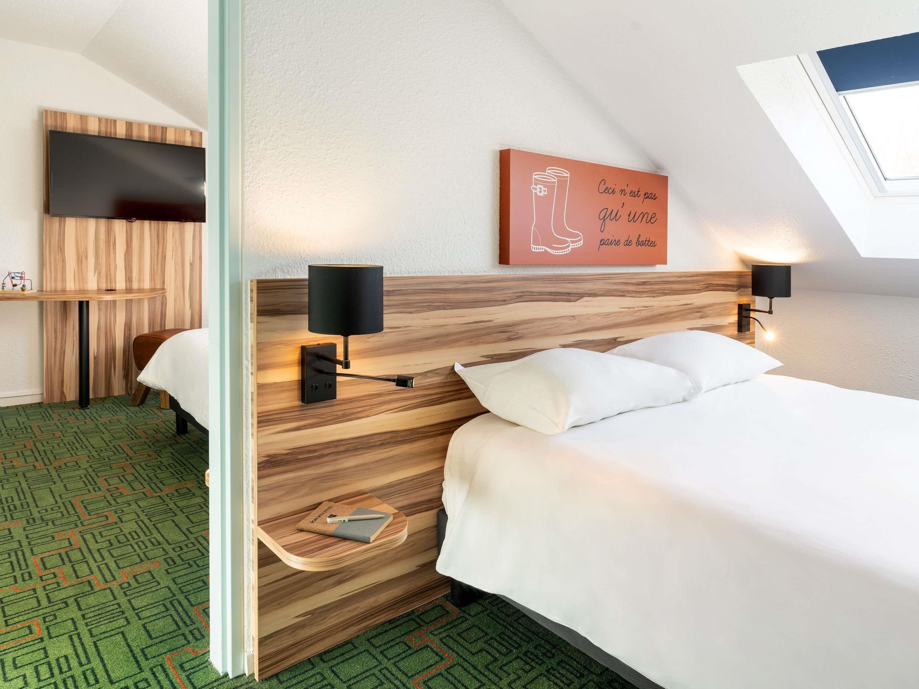 A modern hotel room with a wooden headboard, two bedside lamps, a flat-screen TV, a small table with a notebook, and a green patterned carpet.