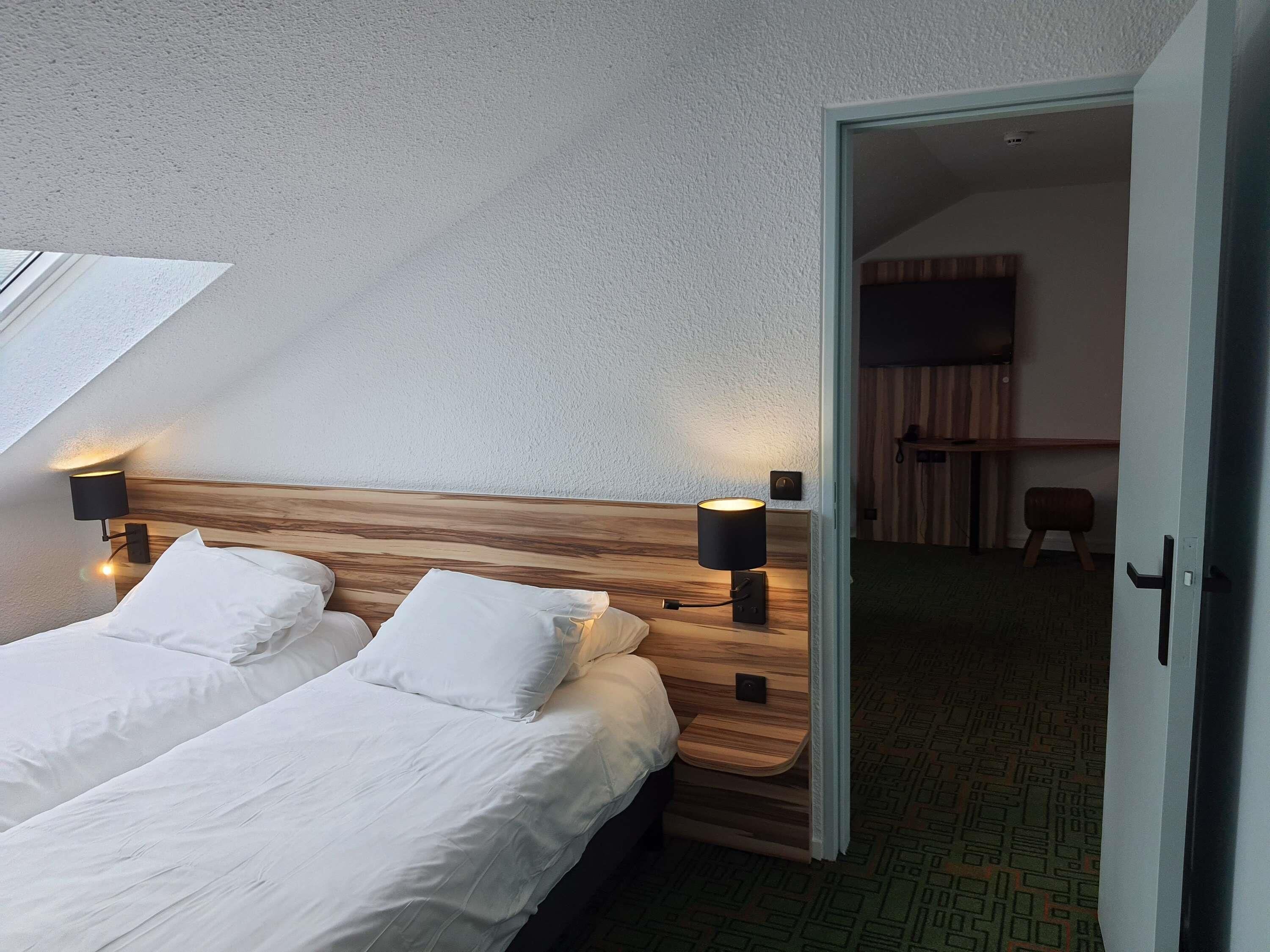 Camera d'hotel con testiera in legno, due letti bianchi, due lampade da comodino e un lucernario.