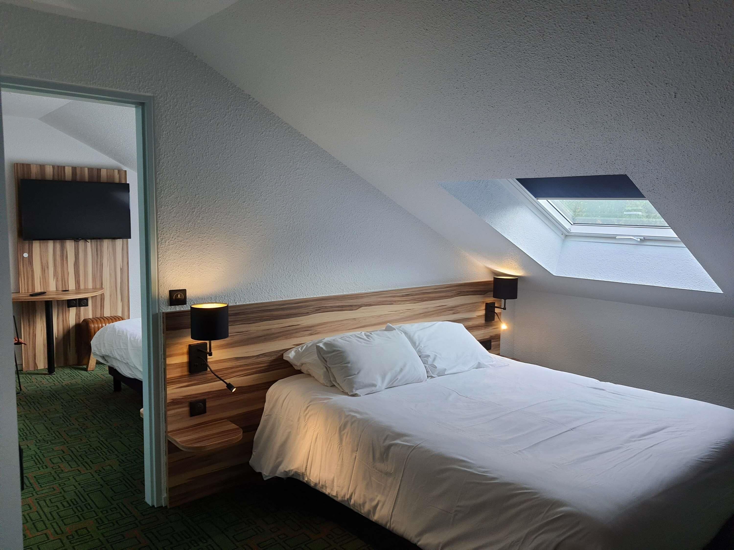 Un dormitorio con techo inclinado, tragaluz, cama con ropa de cama blanca, cabecero de madera y televisor montado en la pared.
