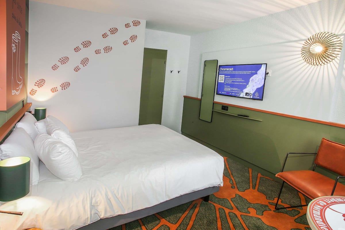 A hotel room with a bed, a television mounted on the wall, a chair, and a carpet with a geometric pattern.