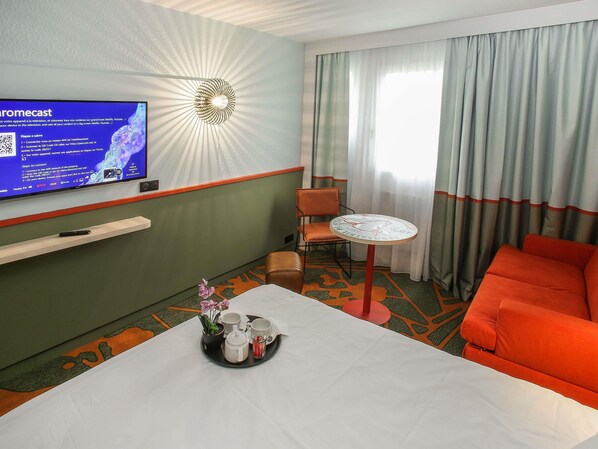 Habitación de hotel con televisor de pantalla plana montado en la pared, una mesita con una bandeja de tazas y una flor, un sofá rojo y una ventana con cortinas.