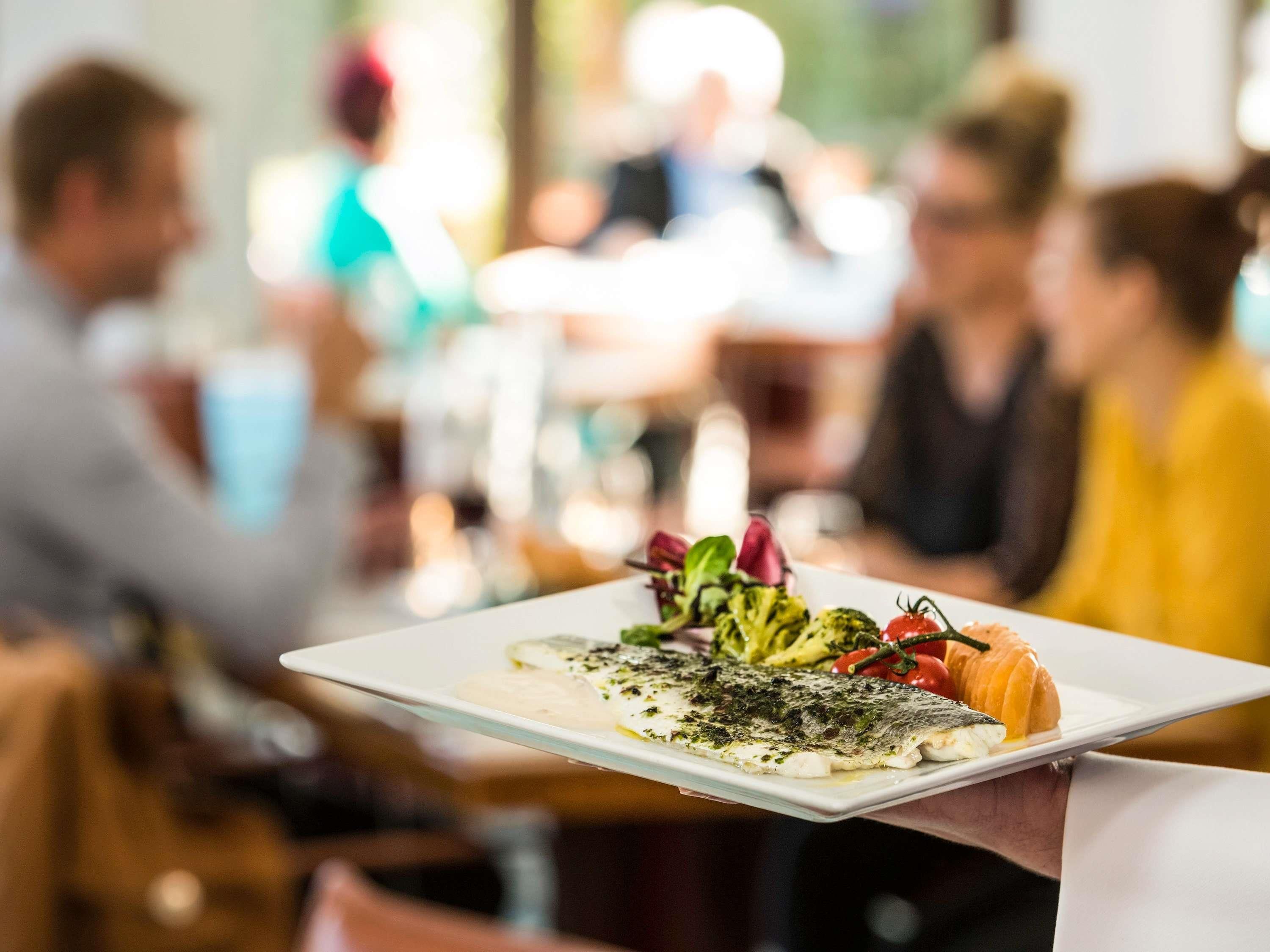 En person håller i en tallrik med en fiskrätt, grönsaker och en brödskiva, och bilden är tagen i en restaurangmiljö med andra gäster i bakgrunden.