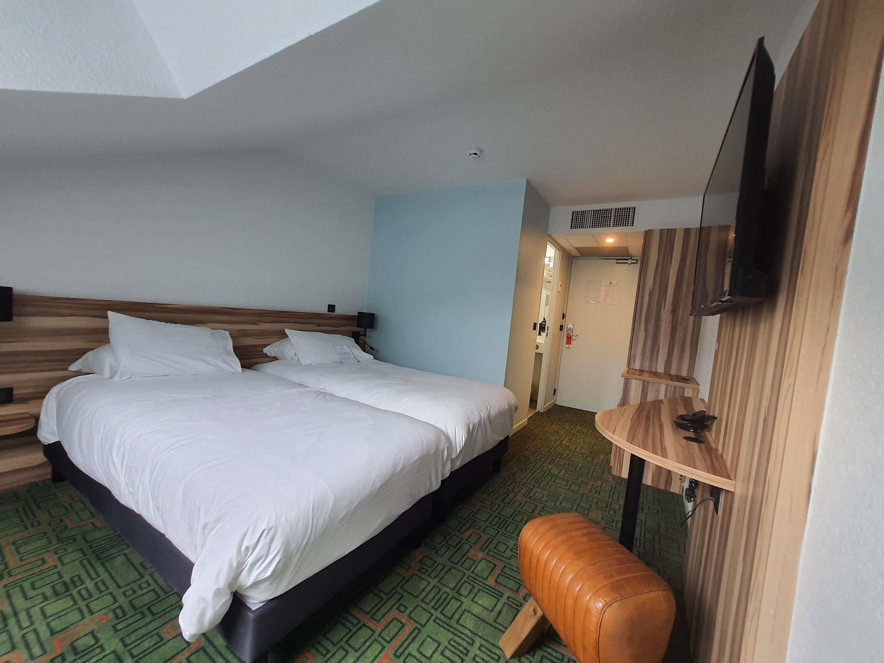 Habitación de hotel con una cama grande, un pequeño banco, un televisor montado en la pared y una puerta que da a un baño.