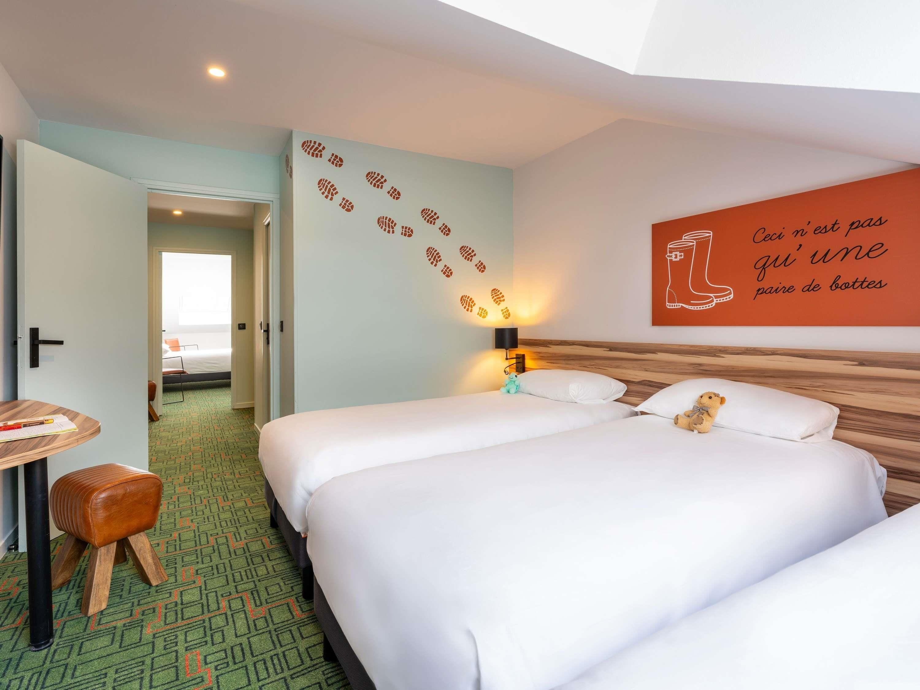 A hotel room with two beds, a desk with a chair, a wooden stool, a wall with a shoe pattern, and a wall with a boot illustration.