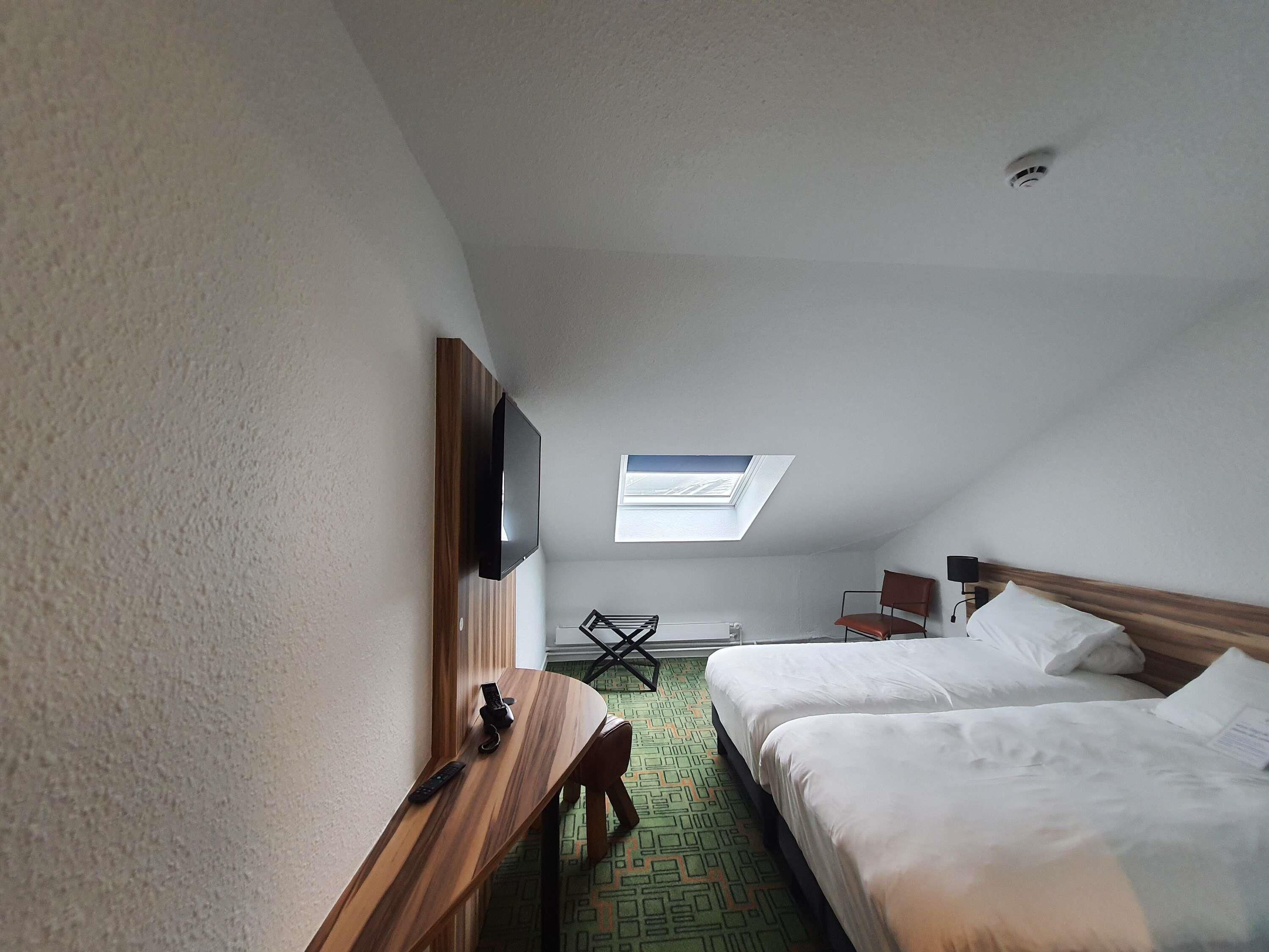 A hotel room with a TV mounted on the wall, a desk with a chair, two beds, a skylight, and a ceiling-mounted smoke detector.