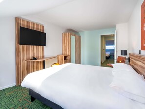 Et hotelværelse med en stor seng, et TV ophængt på en træpanel, et skrivebord med en stol, og udsigt til et andet værelse gennem en åben dør.