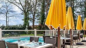Een zwembadterras met rieten stoelen en tafels, gele parasols en een helder uitzicht op het zwembad.