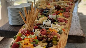 Une longue planche en bois, garnie de différents fromages, de charcuteries et de fruits, est disposée sur une table sous une tente.
