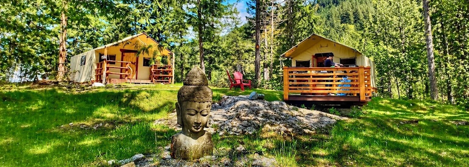 A rustic cabin with a wooden deck, surrounded by lush greenery and a Buddha statue in the foreground.