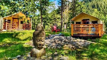 A rustic cabin with a wooden deck, surrounded by lush greenery and a Buddha statue in the foreground.