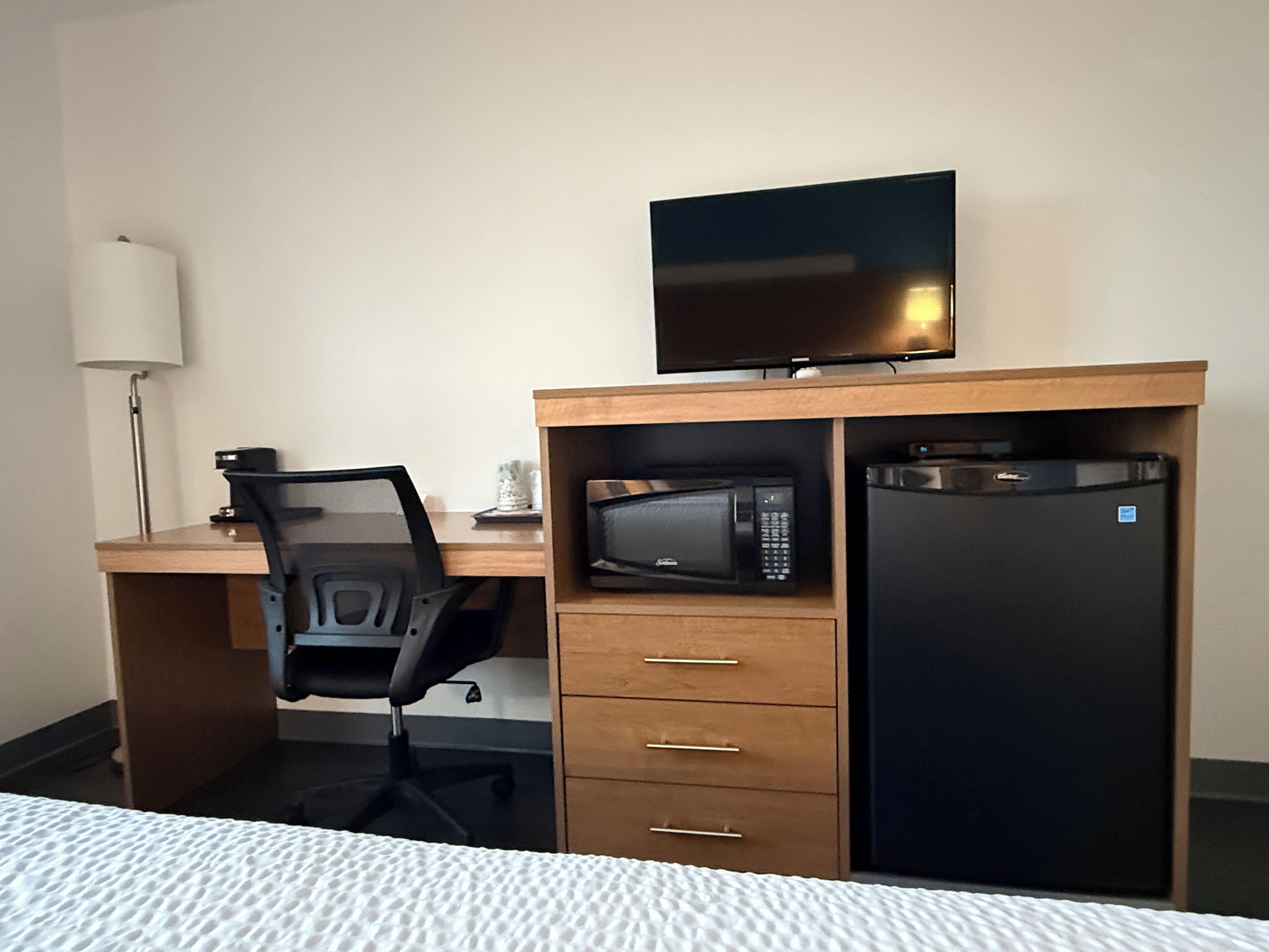 A hotel room with a wooden desk, a black office chair, a flat-screen TV, a microwave, and a refrigerator.