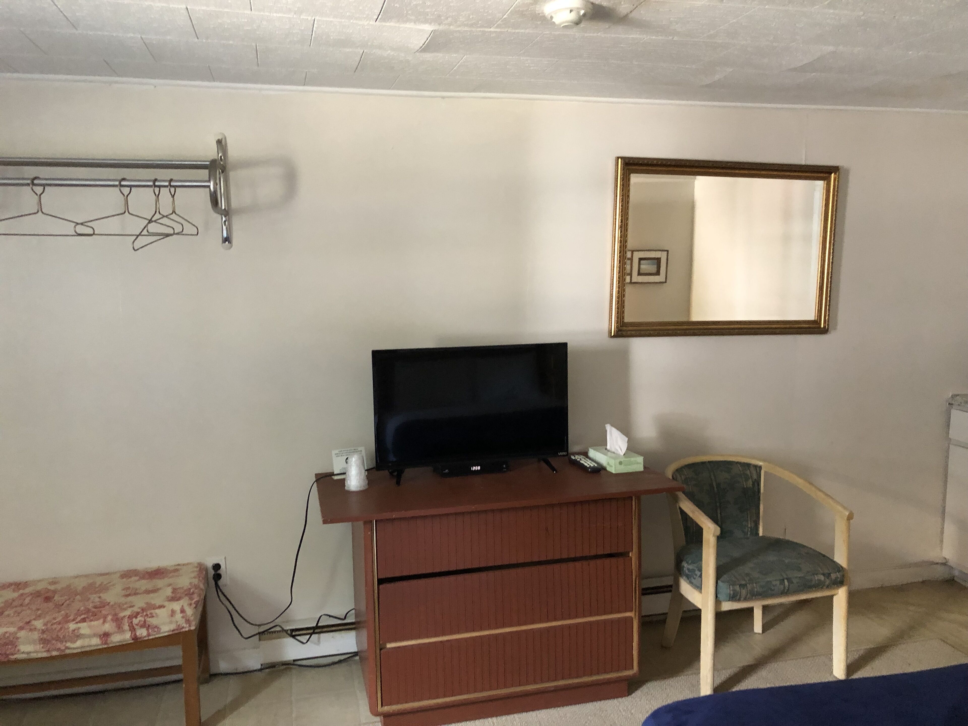 Ein Zimmer mit Fernseher auf einer Holzkommode, einem Sessel, einer Bank und einem Kleiderständer mit Kleiderbügeln.