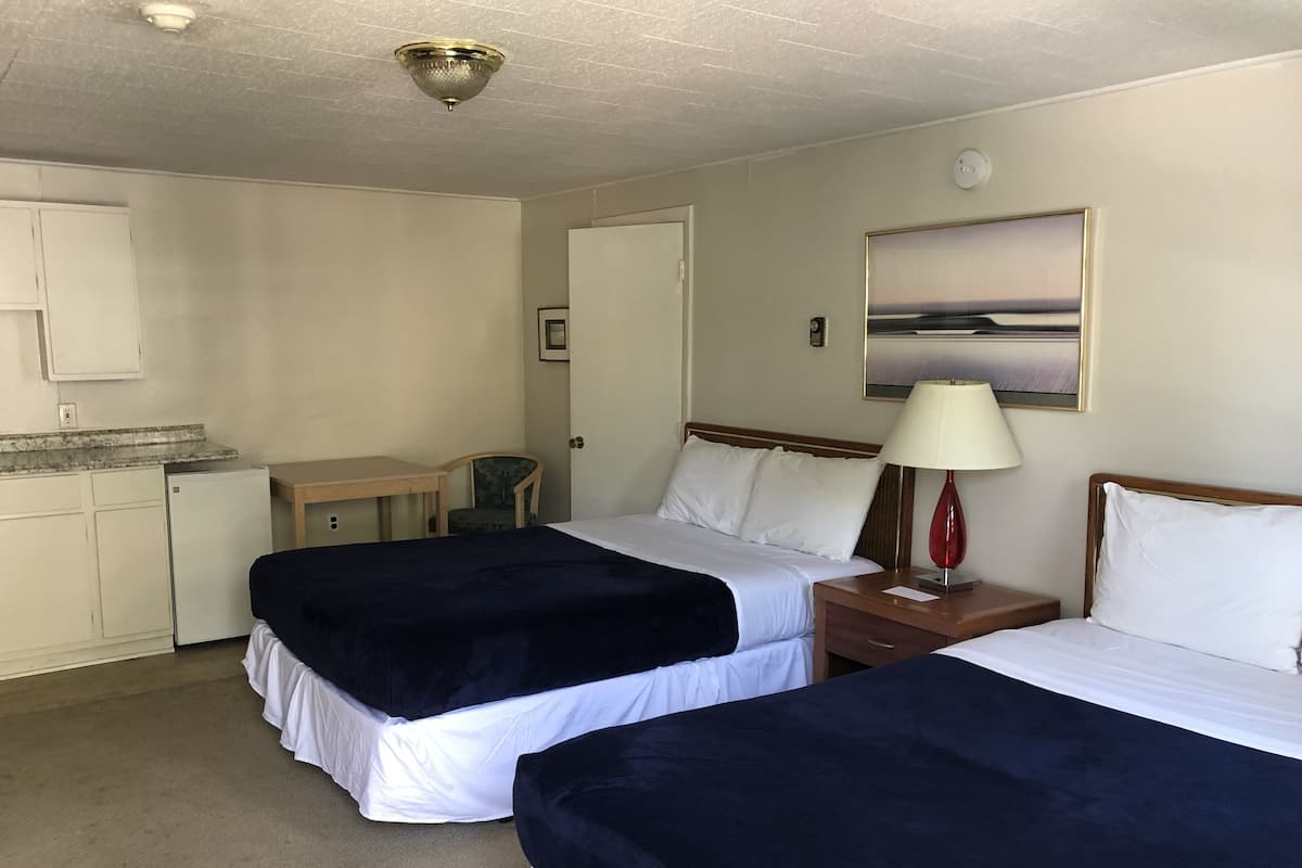 A hotel room with two beds, a desk, a chair, a small kitchenette, and a painting on the wall.